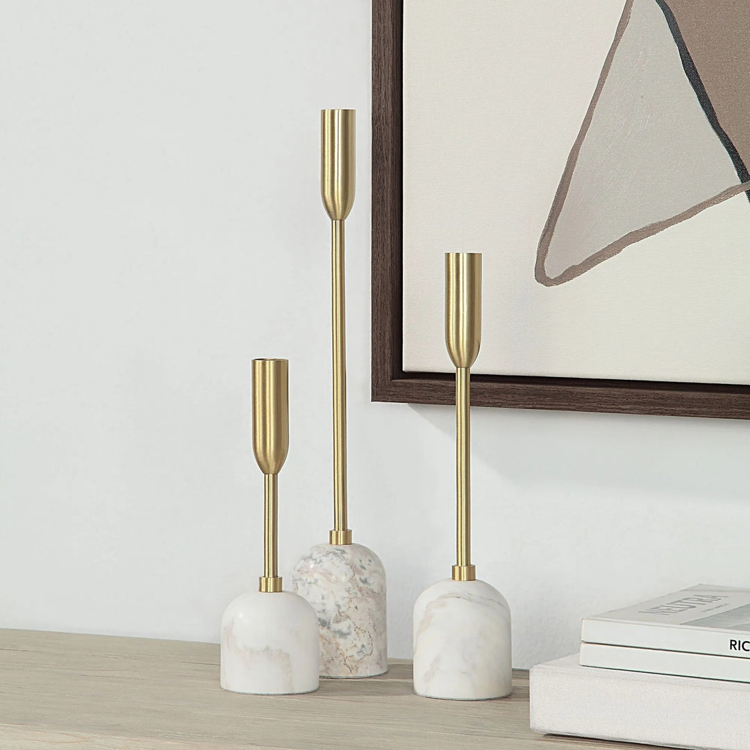 Three white and gold candle holders are arranged on a surface, with a framed artwork visible in the background.