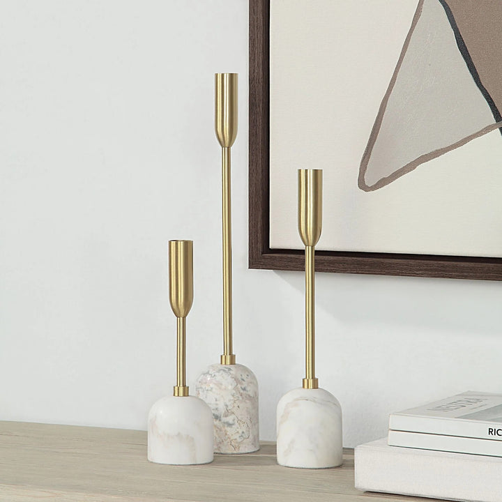 Three white and gold candle holders are arranged on a surface, with a framed artwork visible in the background.