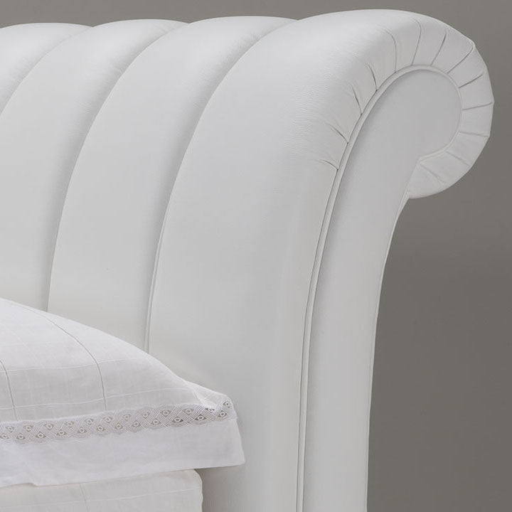 Close-up of a white leather headboard with a curved, rolled top and vertical channel tufting. A white pillow with lace trim rests on the bed.