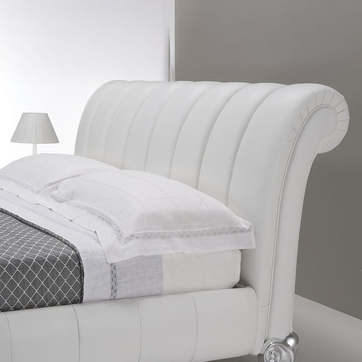 A white leather bed frame with a curved headboard, featuring channel tufting. The bed is made up with white linens, including pillows and a grey patterned quilt, and is set against a neutral background. A lamp is partially visible on the left.