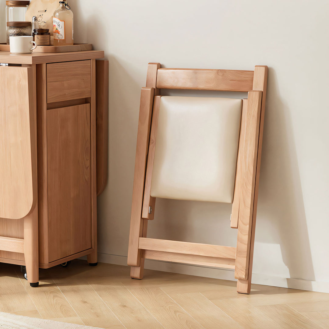 Light wood folding chair with a cream upholstered seat leaning against a wall next to a wooden cabinet.