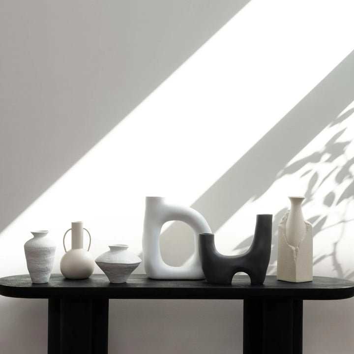 Collection of ceramic vases in various shapes and neutral colors displayed on a black table against a white background with natural light and shadows.