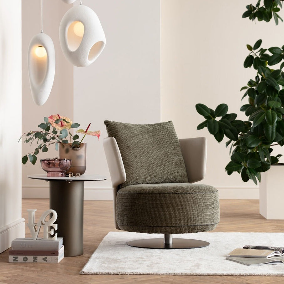 An accent chair with a green fabric seat and back, cream-colored armrests, set in a room setting with decorative lighting, a side table with a vase of flowers, and a large plant.