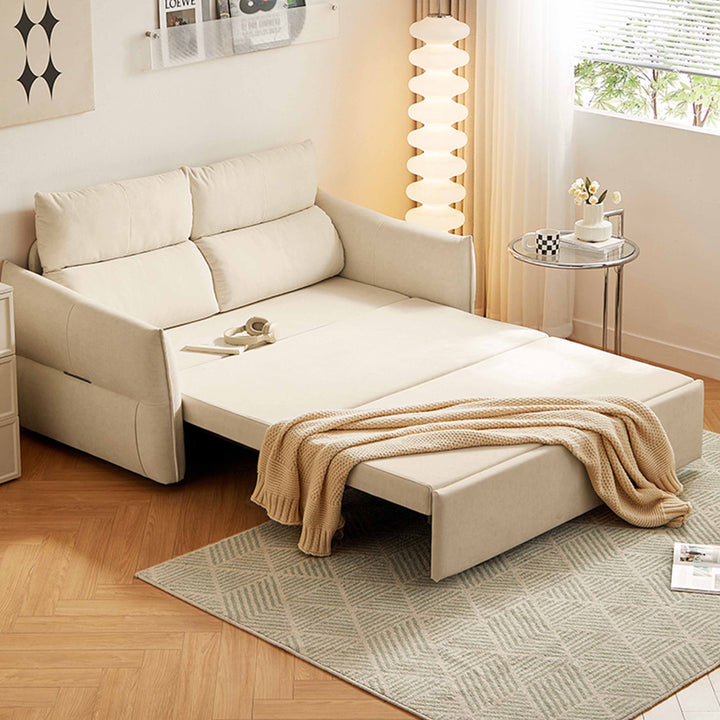 A sofa bed in a room setting with a rug, side table, and lamp. The sofa is beige and has been partially opened. A blanket and some accessories are placed on it.