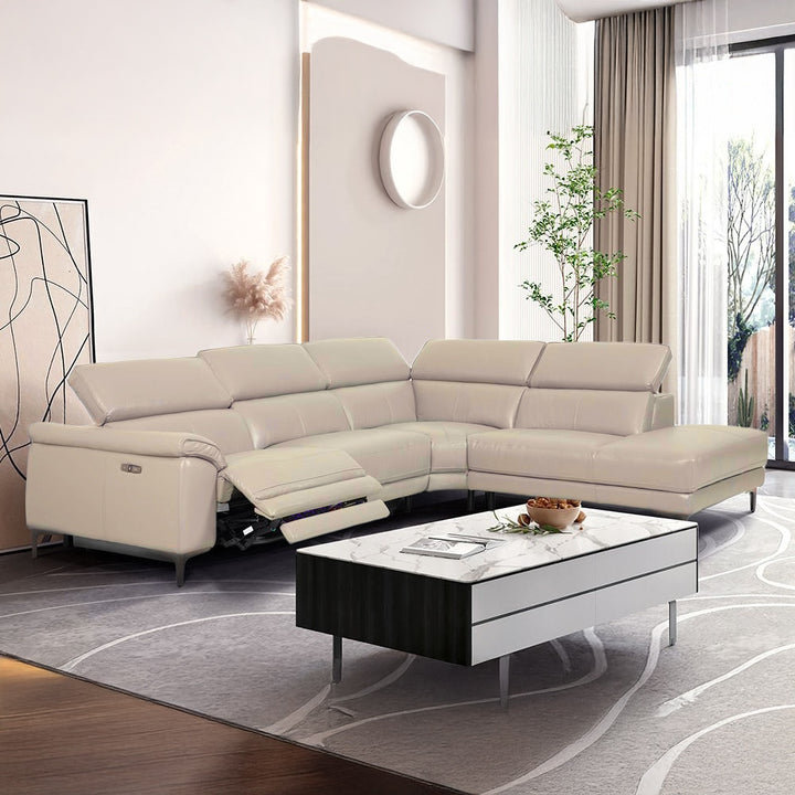 A luxurious cream-colored leather sectional sofa with reclining seats is arranged in a modern living room setting. The sofa faces a contemporary coffee table with a marble top and dark wood base. The room features a large abstract artwork on the wall, a potted plant, and sheer curtains allowing natural light to enter.
