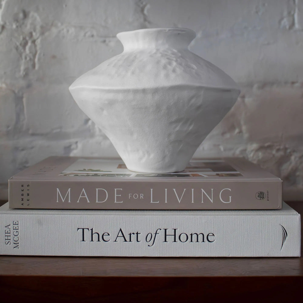 A white ceramic vase rests on top of two books, the background features a white brick wall, styled for home decor.