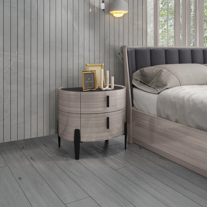 A grey, two-drawer nightstand with a wood grain finish stands beside a bed in a room with grey plank walls and grey wood floors. Decorative items, including picture frames and candlesticks, sit on top of the nightstand.