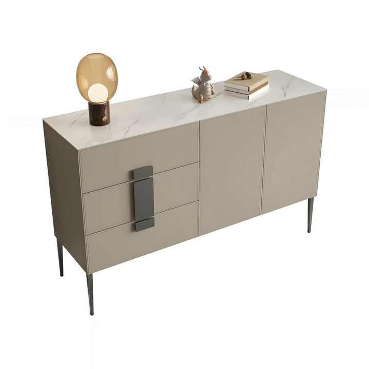 A light beige sideboard with a white marble-effect ceramic top, featuring three drawers with dark grey rectangular handles and two cabinet doors. The sideboard is supported by thin black metal legs and is accessorized with a modern amber glass lamp and a small decorative dragon figurine with stacked books on top.
