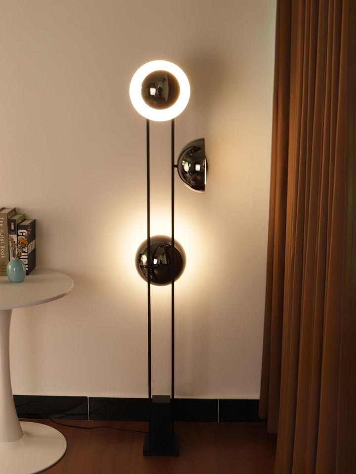 A modern floor lamp with a black metal frame and spherical glass shades is placed in a room setting with a table, books, and curtains.