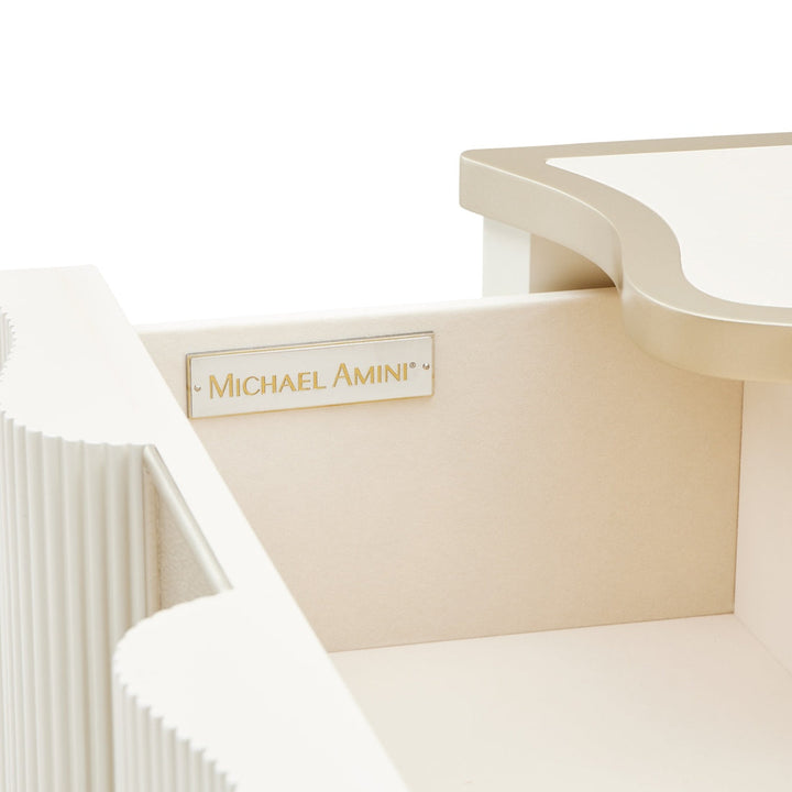 A close-up studio shot of a Michael Amini nightstand drawer on a white background, showcasing a creamy beige color palette and fluted detailing.