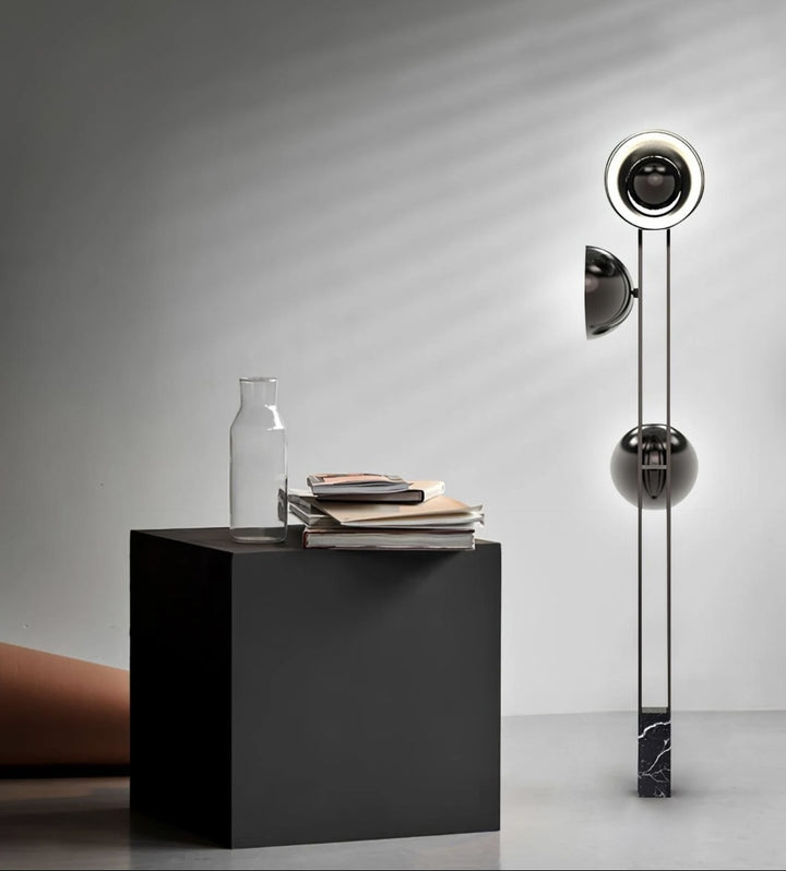 A modern floor lamp with spherical shades and a black marble base stands next to a black cube table with books and a glass bottle, set against a minimalist gray backdrop.