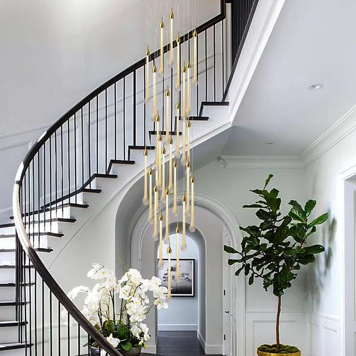 A modern chandelier hangs in a hallway with a curved staircase and plants.