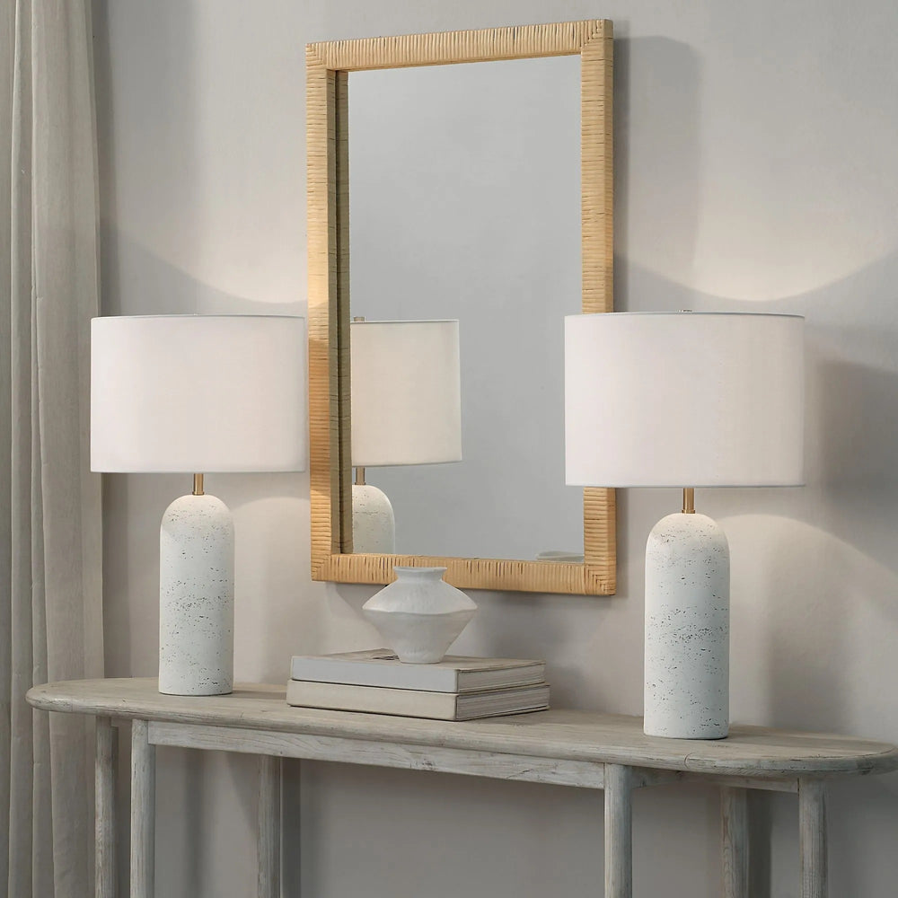 A scene featuring two marble lamps with white shades on a console table, complemented by a rattan-framed mirror and decorative items.