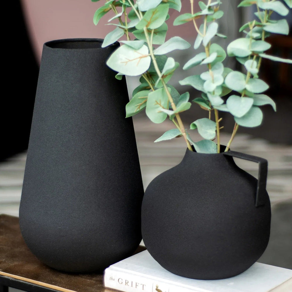 Two black iron vases, one tall and tapered, the other round with a handle, sitting on a table with a book and eucalyptus branches in a room setting.