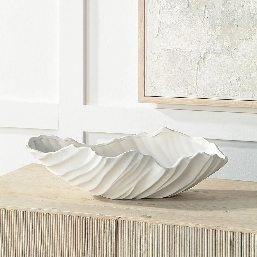 A white ceramic vase with a shell-like, ridged design sits atop a light wood cabinet in a room setting, with a framed artwork visible in the background.