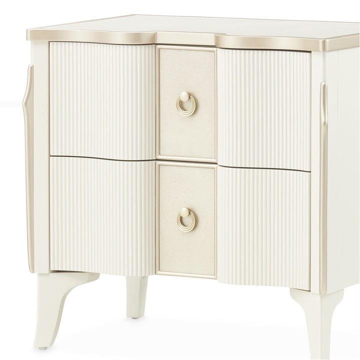 A two-drawer cream-colored nightstand with gold accents, featuring reeded drawer fronts and ring pulls, set against a white background.