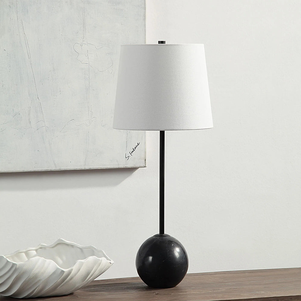 The image shows a lamp featuring a white lampshade, a black steel stem, and a black marble base displayed on a wooden surface in a room setting.