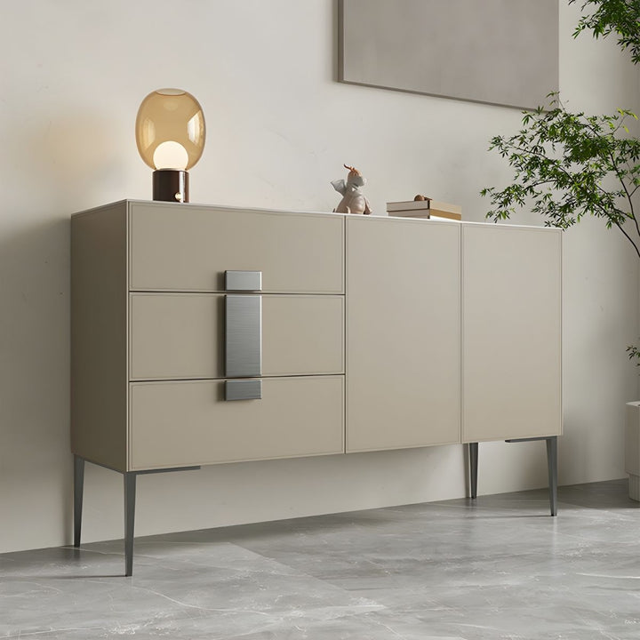 A light taupe sideboard with three drawers and two cabinet doors, accented with sleek champagne gold handles. The sideboard is positioned in a well-lit room with a marble-effect floor, a table lamp with a glass shade on top, and a potted plant in the background.