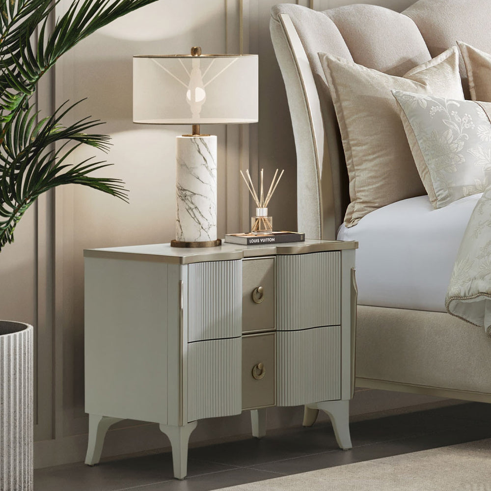 A bedside table with two drawers is placed next to a bed, featuring a lamp and decorative items on top, in a room setting.